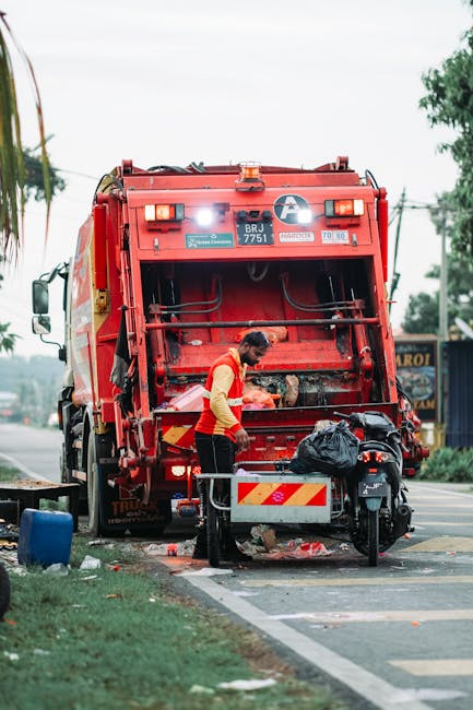 A worker wearing a red and yellow uniform is standing beside a large red rubbish collection truck on a roadside. The truck has a metallic, textured finish with visible hydraulic components and safety 